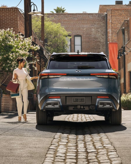 A woman in white with shopping bags reaches for the open tailgate of a dark blue Infiniti SUV on a cobblestone street.