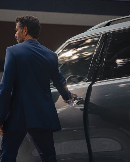 Man in a blue suit opening the driver's side door of a dark-colored car.