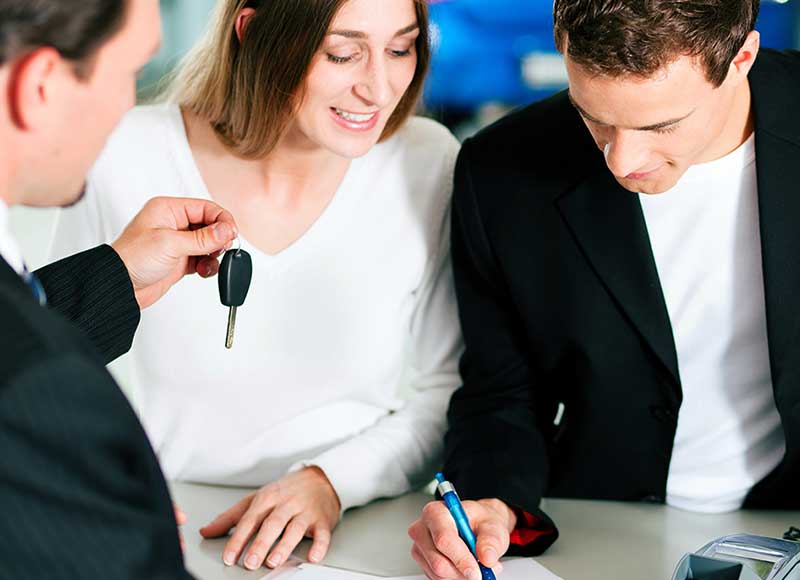 A couple signs paperwork while a salesperson hands them car keys.