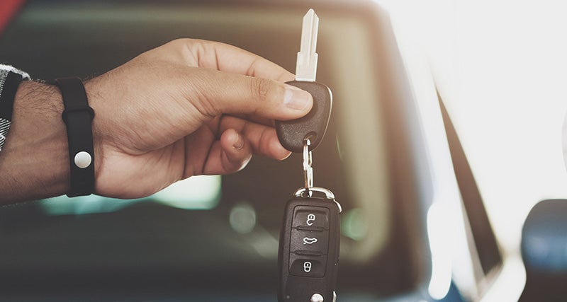 A hand holds a set of car keys with a key fob in front of a blurred vehicle background