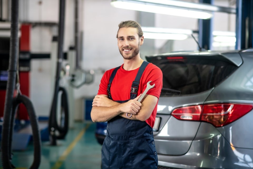 A person in a workshop uniform stands with arms crossed holding a wrench in front of a car