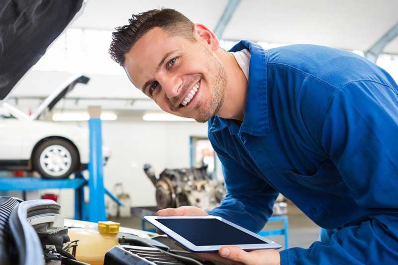 A person in a blue work uniform leans over a car engine while holding a tablet in an auto repair shop